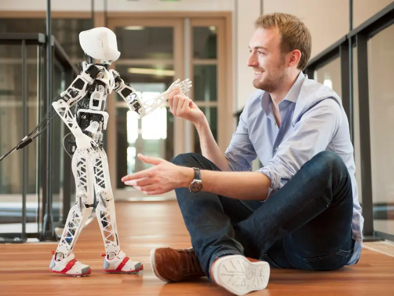 A researcher sitting on the floor, face to face with Poppy, a small white 3D-printed humanoid robot, their hands almost touching.