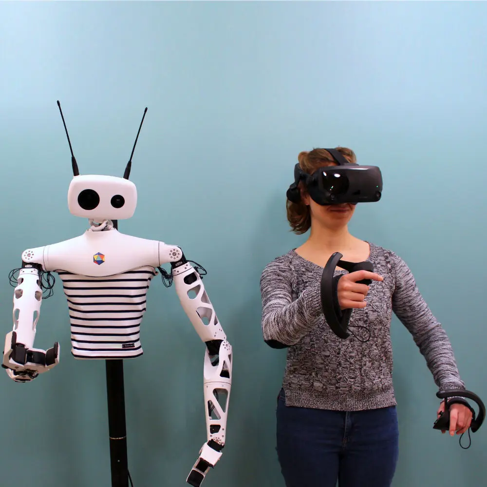 Reachy standing next to a person wearing a VR headset and holding two controllers, both raising an arm in the same pose.
