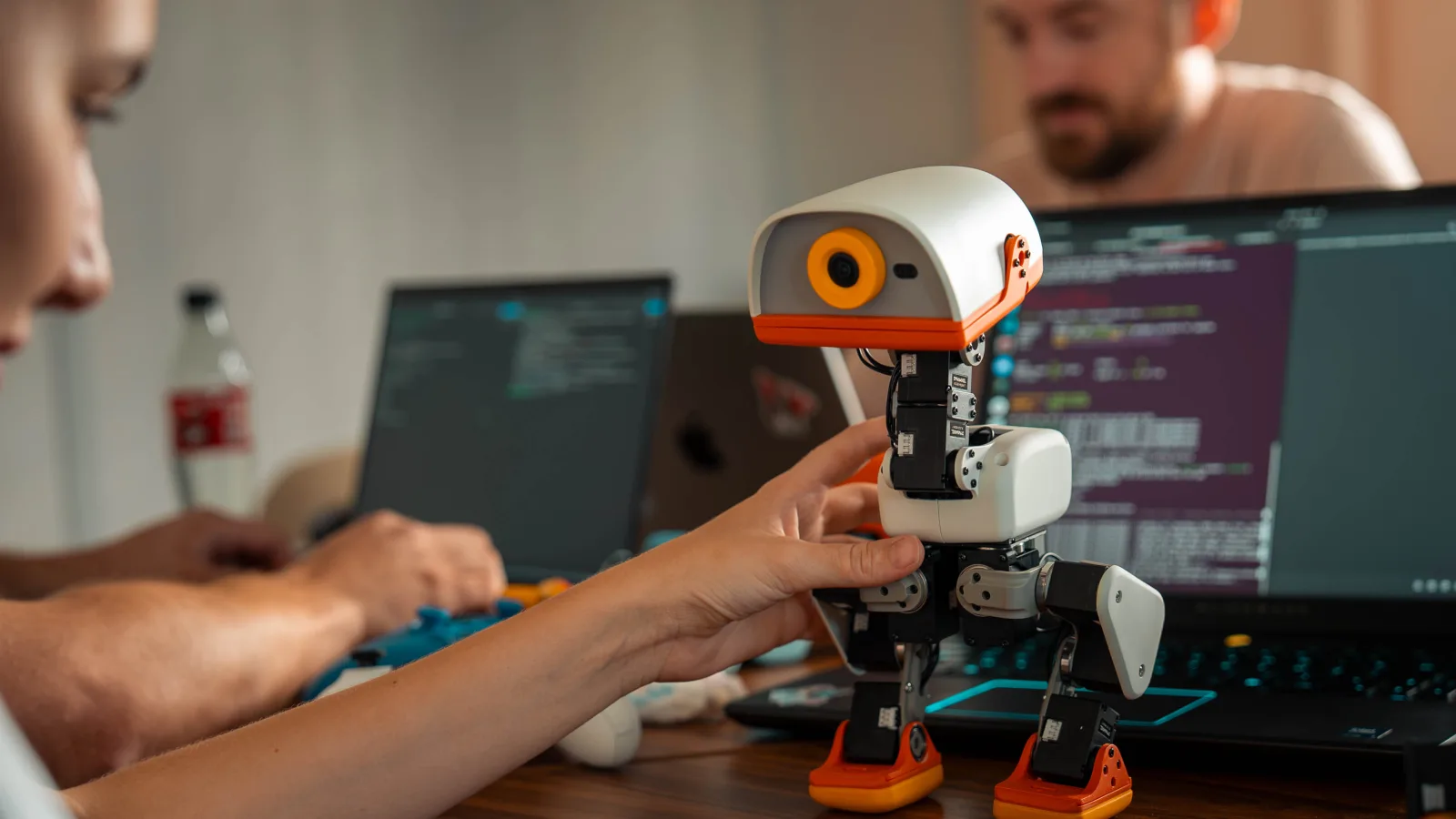 A hand holding a white-and-orange Microduck upright on a desk, between two laptops running code