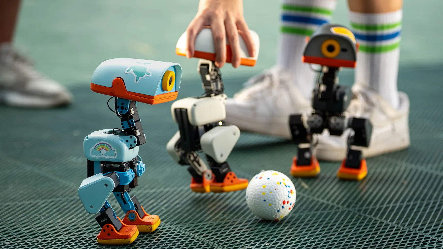 Three Microducks around a ball on a turf pitch, a hand steadying one of them